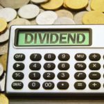 dividend stocks: A calculator projecting the word "DIVIDEND" rests on a pile of gold and silver coins.
