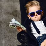 A photo of a young boy wearing sunglasses, jeans, a blazer, a white shirt and suspenders holding money in various denominations in one hand and sitting in a plush chair.
